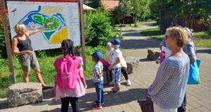 Frauen und Kinder stehen an einer Ansichtstafel und hören einer Frau zu, die etwas dazu erklärt.