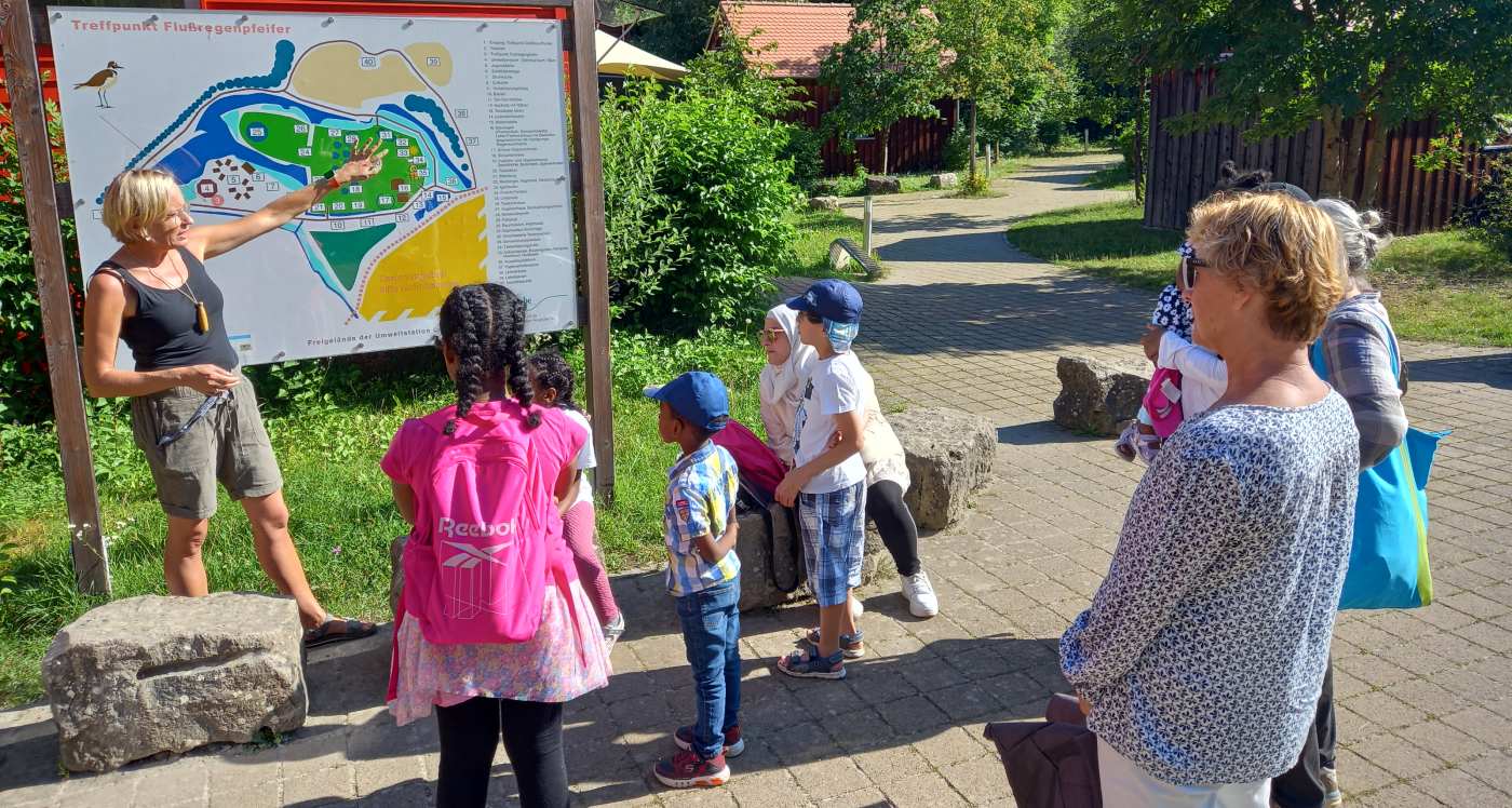 Frauen und Kinder stehen an einer Ansichtstafel und hören einer Frau zu, die etwas dazu erklärt.